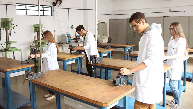 Estudantes de jaleco em aula prática em oficina com bancadas de trabalho